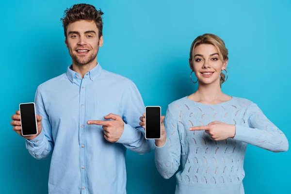 Sonriente hombre y mujer señalando con los dedos a los teléfonos inteligentes con pantallas en blanco sobre fondo azul - foto de stock