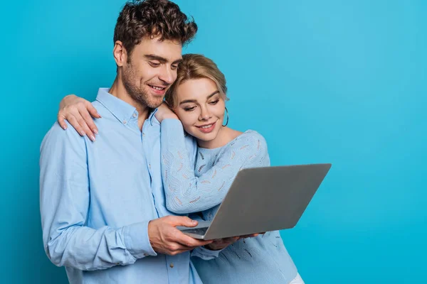 Felice ragazza abbracciando fidanzato sorridente mentre guardando il computer portatile insieme su sfondo blu — Foto stock