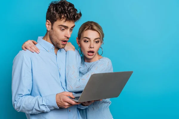 Schockiertes Mädchen umarmt überraschten Freund, während sie gemeinsam auf Laptop auf blauem Hintergrund — Stockfoto