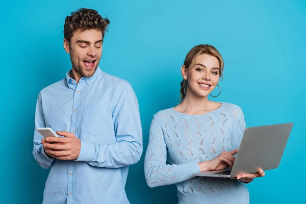 Hombre alegre charlando en el teléfono inteligente y mirando a la novia con el ordenador portátil en el fondo azul - foto de stock