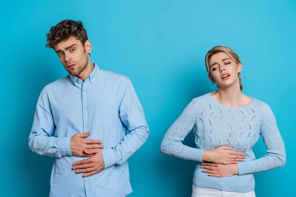 Perturbado homem e mulher tocando estômagos enquanto sofre de dor abdominal no fundo azul — Fotografia de Stock