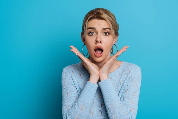 Scioccato ragazza con bocca aperta che tiene le mani vicino al viso mentre guardando la fotocamera su sfondo blu — Foto stock