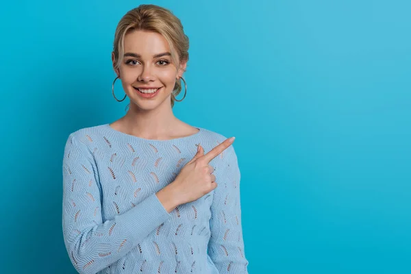 Menina bonita sorrindo para a câmera ao apontar com o dedo no fundo azul — Fotografia de Stock