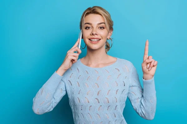 Positive girl showing idea sign while talking on smartphone on blue background — Stock Photo