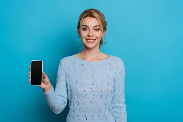 Весела дівчина посміхається на камеру, показуючи смартфон з порожнім екраном на синьому фоні — стокове фото