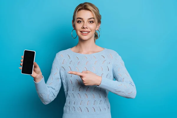 Улыбающаяся девушка указывает пальцем на смартфон с пустым экраном на синем фоне — стоковое фото