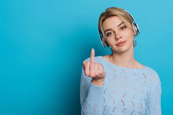 Ragazza scettica mostrando dito medio durante l'ascolto di musica in cuffie wireless su sfondo blu — Foto stock