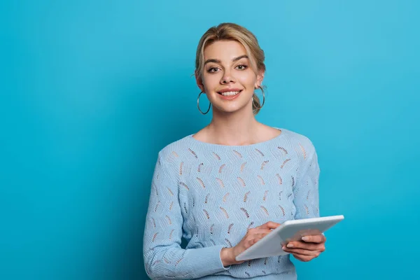 Fröhliches Mädchen lächelt in die Kamera, während es ein digitales Tablet auf blauem Hintergrund benutzt — Stockfoto