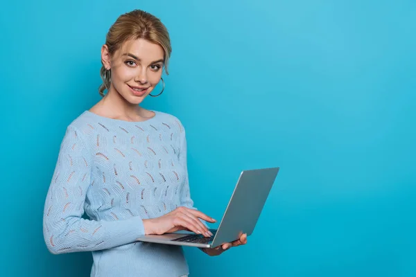 Fröhliches Mädchen lächelt in die Kamera, während es Laptop auf blauem Hintergrund benutzt — Stockfoto