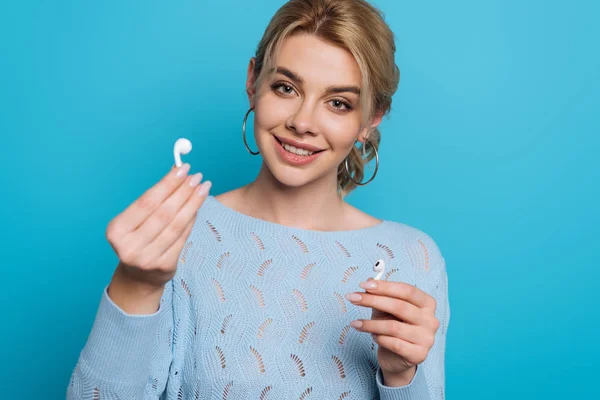 Cheerful girl showing wireless earphones while smiling at camera on blue background — Stock Photo