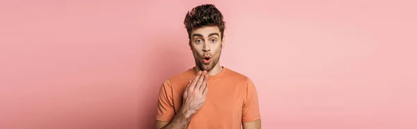 Panoramic shot of shocked young man holding hand near mouth while looking at camera on pink background — Stock Photo