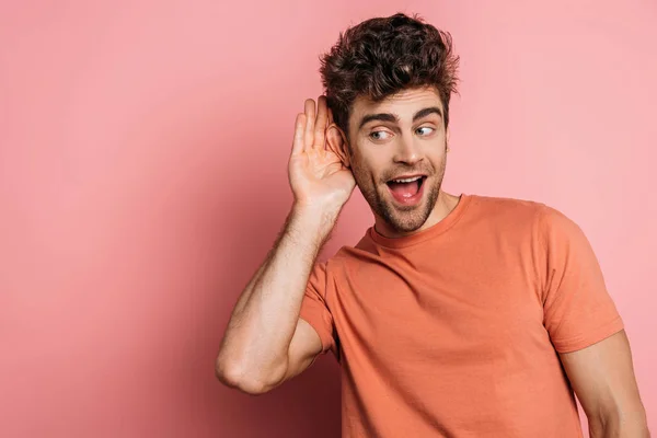 Neugieriger junger Mann belauscht, während er die Hand vor dem Ohr auf rosa Hintergrund hält — Stockfoto