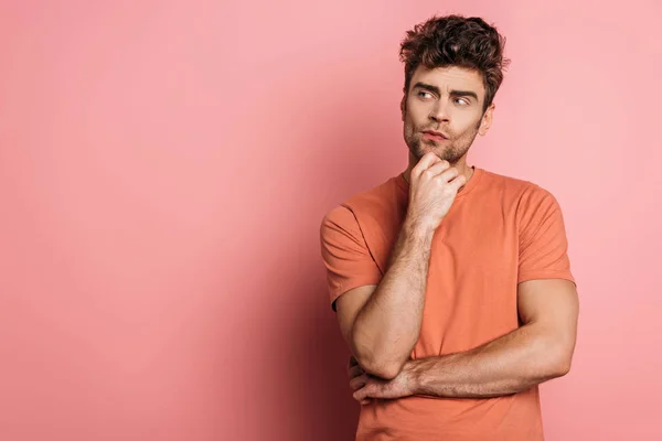 Nachdenklicher junger Mann schaut weg, während er sein Kinn auf rosa Hintergrund berührt — Stockfoto