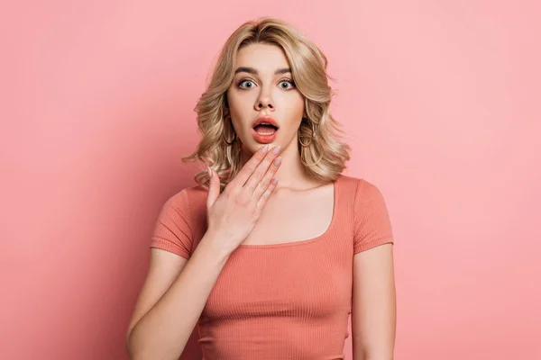 Shocked girl looking at camera while holding hand near open mouth on pink background — Stock Photo