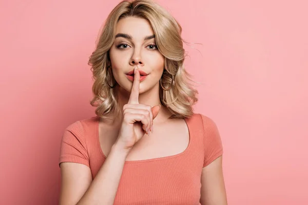 Attractive girl looking at camera while showing hush gesture on pink background — Stock Photo