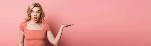 Plano panorámico de chica impactada apuntando con la mano mientras mira a la cámara en el fondo rosa - foto de stock