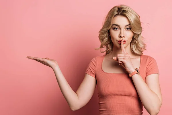 Beautiful girl showing hush gesture and pointing with hand on pink background — Stock Photo