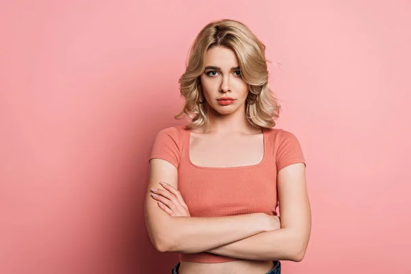 Offended girl looking at camera while standing with crossed arms on pink background — Stock Photo