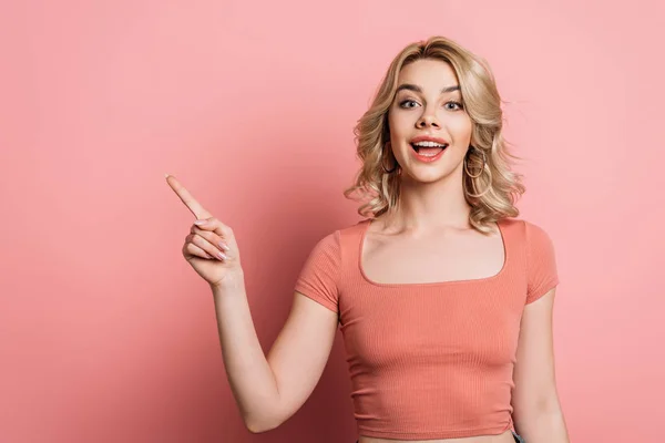 Fröhliches Mädchen zeigt mit dem Finger und blickt in die Kamera auf rosa Hintergrund — Stockfoto