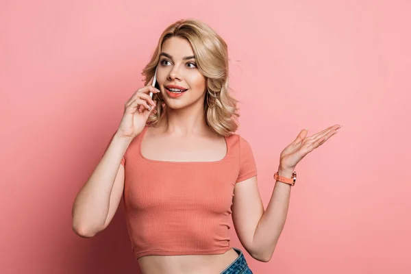 Chica positiva de pie con el brazo abierto mientras habla en el teléfono inteligente sobre fondo rosa - foto de stock