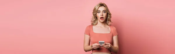 Plano panorámico de chica sorprendida mirando a la cámara mientras chatea en el teléfono inteligente sobre fondo rosa - foto de stock