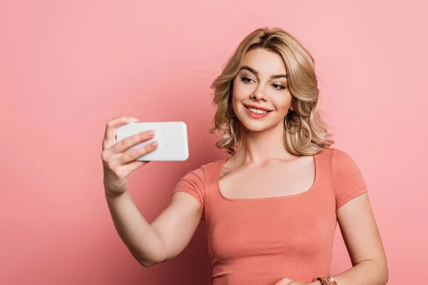 Chica atractiva, alegre tomando selfie en el teléfono inteligente sobre fondo rosa - foto de stock