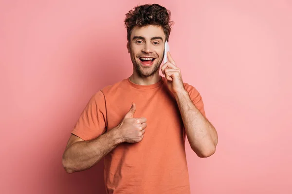 Happy young man showing thumb up while talking on smartphone on pink background — Stock Photo