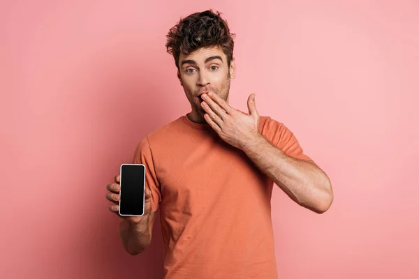 Шокированный молодой человек, прикрывающий рот рукой, показывая смартфон с пустым экраном на розовом фоне — стоковое фото