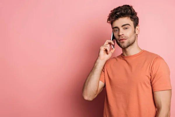 Serious young man looking at camera while talking on smartphone on pink background — Stock Photo