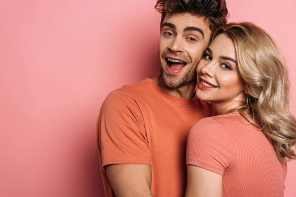 Feliz jovem casal abraçando enquanto sorrindo para a câmera no fundo rosa — Fotografia de Stock