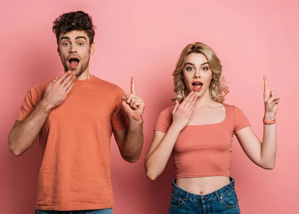 Sorprendido hombre y mujer mostrando signos de idea mientras mira a la cámara en el fondo rosa - foto de stock