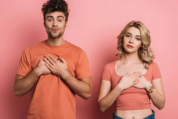 Tricky young man and serious, honest girl holding hands on chest while looking at camera on pink background — Stock Photo