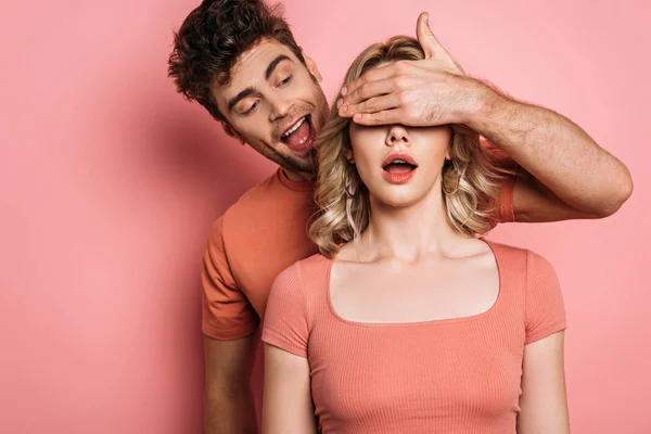 Hombre complicado cubriendo los ojos de la chica sorprendida con la mano sobre fondo rosa - foto de stock