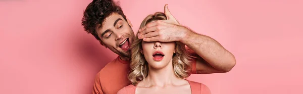 Panoramic shot of tricky man covering eyes of surprised girl with hand on pink background — Stock Photo