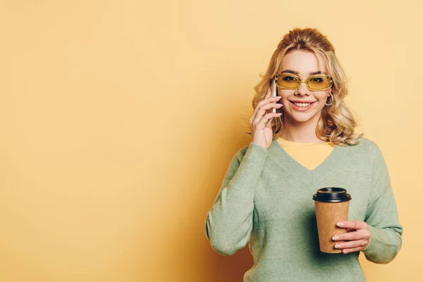 Menina alegre falando no smartphone, segurando café para ir e sorrindo para a câmera no fundo amarelo — Fotografia de Stock