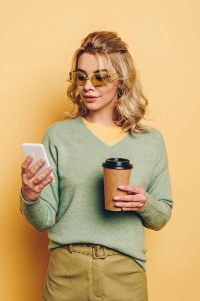 Chica elegante y sonriente sosteniendo café para ir mientras charlas en el teléfono inteligente sobre fondo amarillo - foto de stock