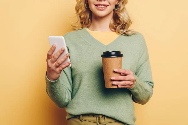 Cropped view of smiling girl using smartphone and holding coffee to go on yellow background — Stock Photo