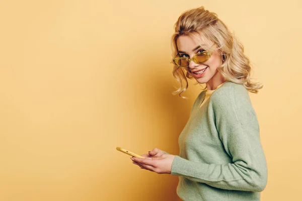 Chica bonita en gafas charlando en el teléfono inteligente y sonriendo a la cámara en el fondo amarillo - foto de stock