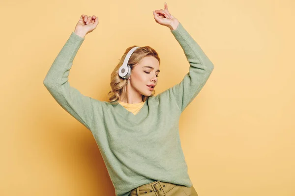 Chica soñadora escuchando música en auriculares inalámbricos y bailando sobre fondo amarillo - foto de stock