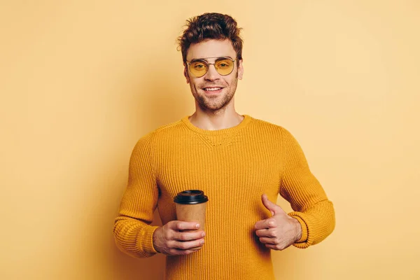 Joven guapo sonriendo a la cámara mientras muestra el pulgar hacia arriba y sosteniendo el café para ir sobre fondo amarillo - foto de stock