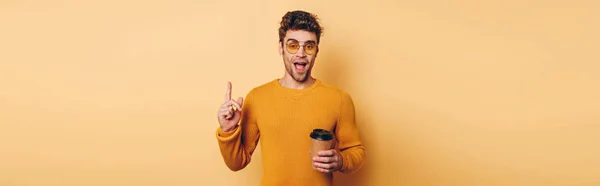 Panoramic shot of excited man showing idea sign while holding disposable cup on yellow background — Stock Photo