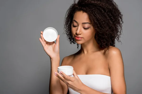 Bela menina afro-americana segurando recipiente de plástico com creme cosmético, isolado em cinza — Fotografia de Stock