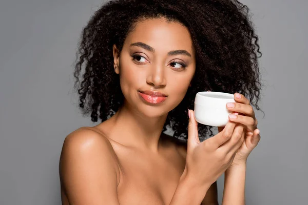 Smiling african american girl holding cosmetic cream, isolated on grey — Stock Photo