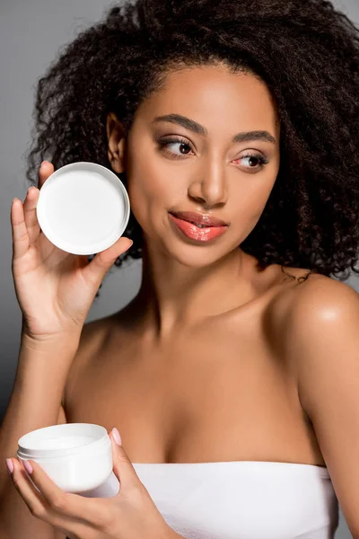 Sorridente menina americana africana segurando recipiente de plástico com creme cosmético, isolado em cinza — Fotografia de Stock