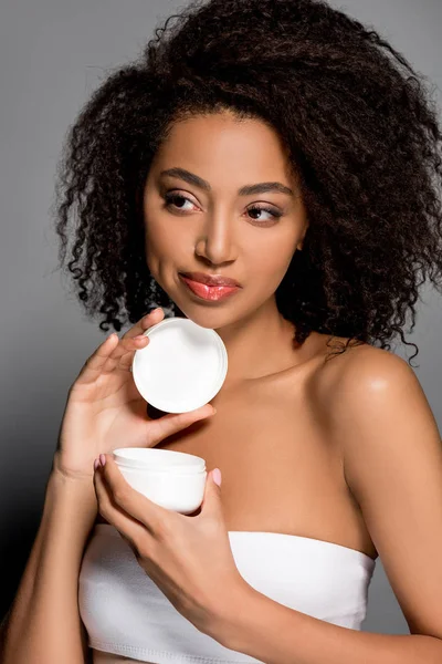 Hermosa chica afroamericana sosteniendo contenedor de plástico con crema cosmética, aislado en gris - foto de stock