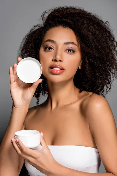 African american girl holding plastic container with cream, isolated on grey — Stock Photo