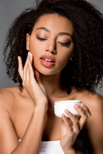 Tierna joven afroamericana mujer aplicando crema facial, aislado en gris - foto de stock