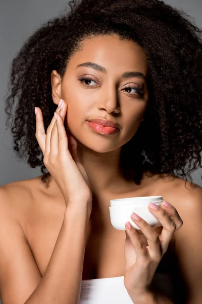 Tierna chica afroamericana aplicando crema facial, aislado en gris - foto de stock