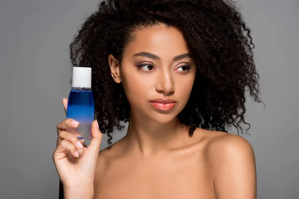 Naked african american girl holding bottle of makeup remover, isolated on grey — Stock Photo