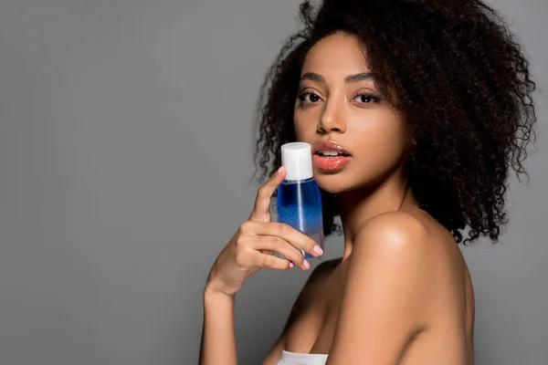 Sexy african american girl holding bottle of makeup remover, isolated on grey — Stock Photo